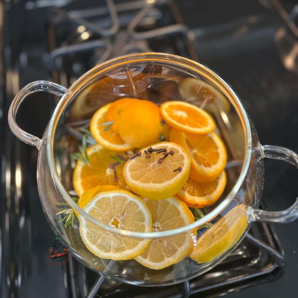 Looking down into a clear pot that is filled with lemon slices, ornage slices, rosemary sprigs, and whole gloves.