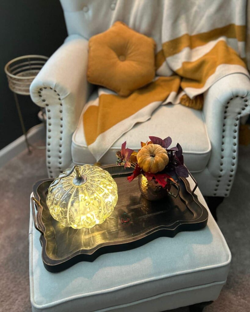 Looking down on a foot stool with a lit up pumpkin and a copper cup with fall leaves in it.