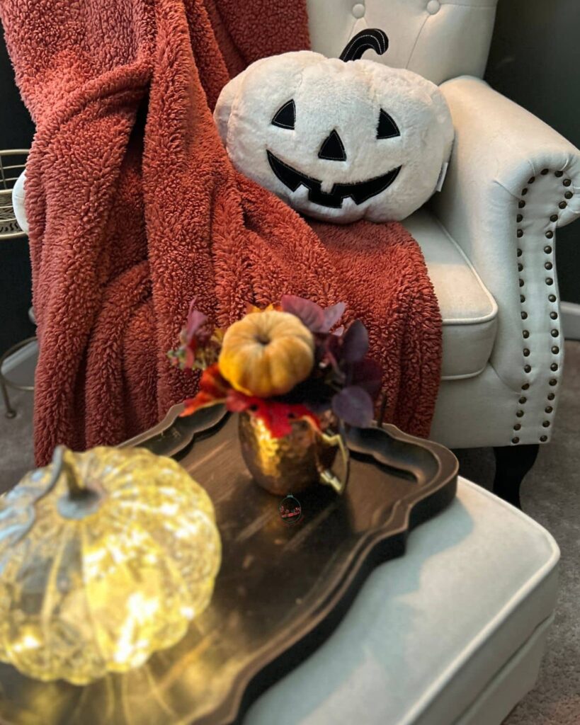 Close up of more cozy fall decor on an foot stoll that has a lit up glass pumpkin.