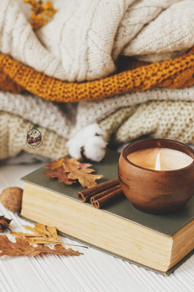 Cozy Halloween colored blankets stacked with a book, candle, cinnamon sticks,and leaves in front of it.