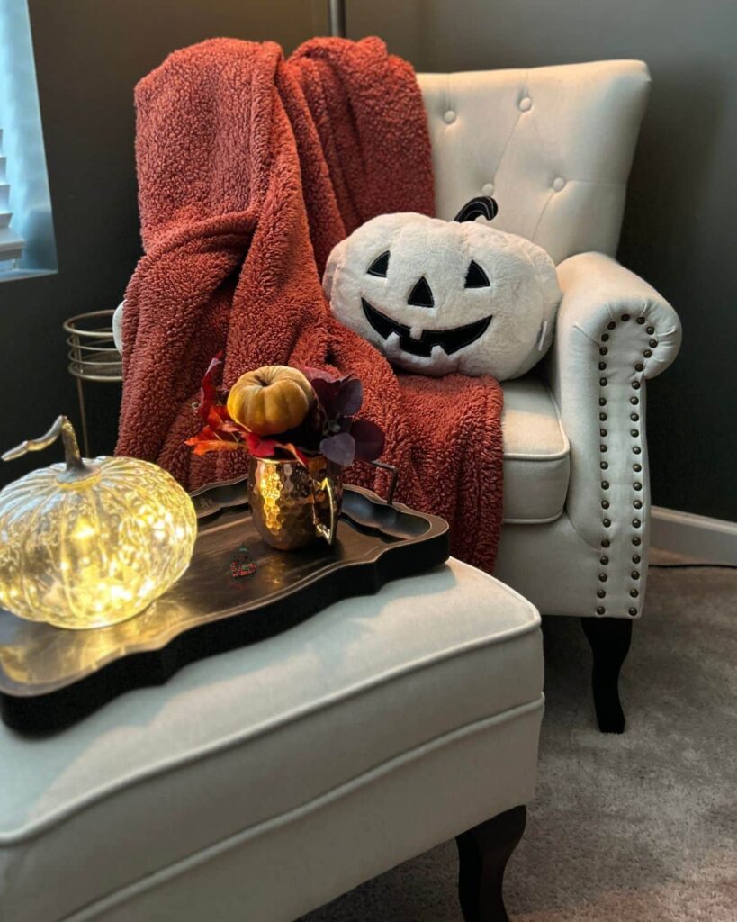 A cozy fall chair that has an orange blanket and large pumpkin pillow on it.