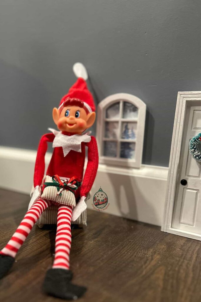 An Elf on the shelf sitting in a cozy miniature chair with a door and window next to him.