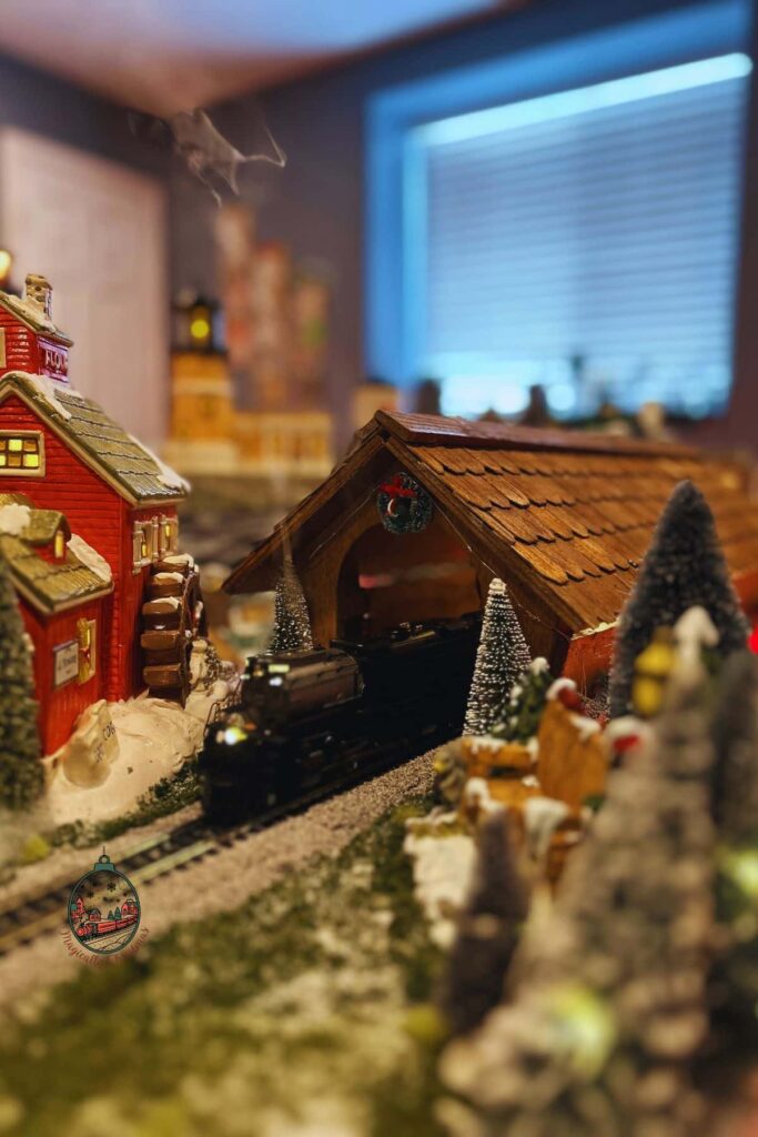 A Broadway Limited Challenger coming out of a wooden bridge on a Christmas village layout.